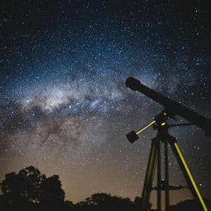 Un telescopio su treppiede si staglia contro un cielo notturno stellato con la Via Lattea sopra la sagoma degli alberi.