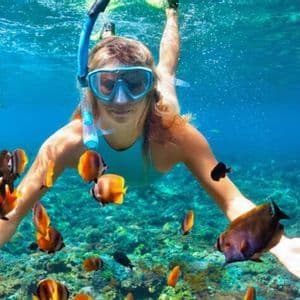 Una donna con maschera da snorkeling nuota sott'acqua, circondata da molti piccoli pesci tropicali colorati sopra una barriera corallina.
