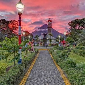 Un sentiero lastricato attraverso un parco lussureggiante conduce a una chiesa con un vulcano sullo sfondo sotto un cielo rosato al tramonto.