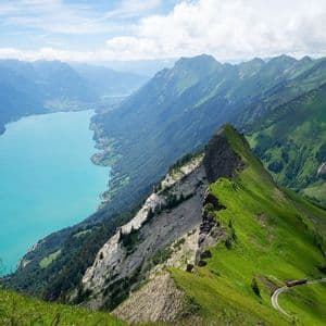 Vue en plongée d'une crête montagneuse verdoyante près d'un lac turquoise, avec un petit train rouge circulant sur une voie.