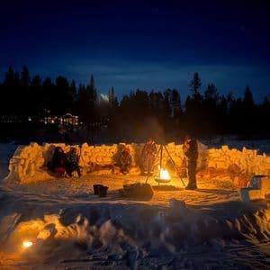 Un viaggio di gruppo WeRoad riunito attorno a un falò di notte in un paesaggio innevato, seduto all'interno di un muro circolare fatto di blocchi di neve.