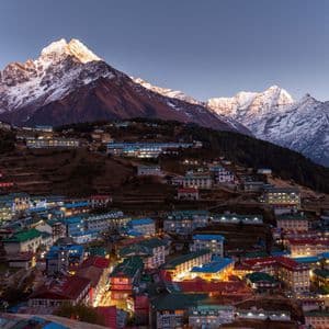Un pueblo iluminado en una ladera aterrazada al anochecer, con grandes montañas nevadas al fondo.