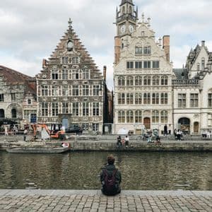 Une personne avec un sac à dos est assise sur un quai pavé, regardant de l'autre côté d'un canal des maisons historiques à pignons sous un ciel nuageux.