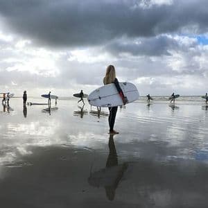 Eine WeRoad-Gruppe von Surfern mit Surfbrettern geht an einem nassen Strand entlang, ihre Spiegelungen sind im Sand sichtbar.