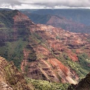 Ein dünner Wasserfall stürzt eine Klippe hinab in eine weite, zerklüftete Schlucht mit geschichteten roten Felsen und grünem Laub.