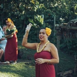 Eine Frau in einem roten Kleid und mit einem Blumen-Lei gestikuliert zu einer sitzenden WeRoad-Gruppenreise in einer üppigen Naturumgebung.