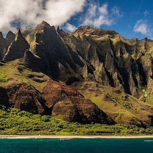 Ein zerklüfteter, grüner Gebirgszug mit scharfen Gipfeln ragt über eine bewaldete Küste, während Boote auf dem blauen Wasser darunter fahren.