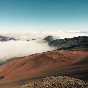 Ein Blick von einem felsigen Berggipfel hinunter auf einen rotbraunen Hang und ein Tal, gefüllt mit einem Wolkenmeer.