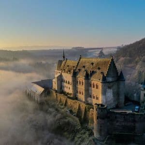 Una veduta aerea di un grande castello in pietra su una collina, che si erge sopra una coltre di nebbia all'alba.