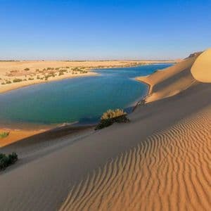 Un lago azzurro forma un'oasi in un vasto deserto, con dune di sabbia dorata e increspate in primo piano sotto un cielo limpido.