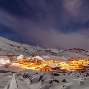 Un villaggio illuminato annidato tra montagne innevate di notte, con un cielo blu scuro e nuvole sopra.