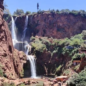 Ein großer, mehrstufiger Wasserfall stürzt rote Felsen hinab in ein Becken, umgeben von üppigem Grün und Gebäuden, die sich in den Hang schmiegen.