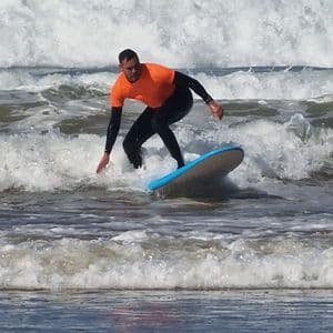 Ein Mann in einem orangefarbenen Hemd und schwarzem Neoprenanzug reitet eine Welle auf einem hellblauen Surfbrett.