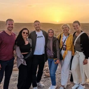 Eine WeRoad Gruppenreise von sechs lächelnden Personen auf einem Hügel mit Blick auf ein Dorf in einer trockenen Landschaft bei Sonnenuntergang.