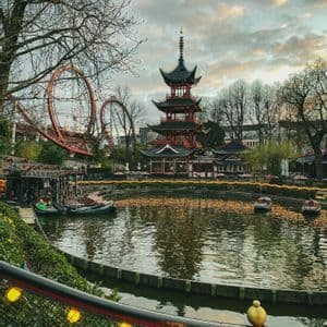 A Japanese pagoda and a red rollercoaster overlook a lake with boats at an amusement park during dusk.