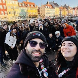 A WeRoad group trip takes a large group selfie on a cobblestone street in front of colorful canal-side buildings.