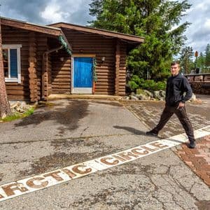 Ein junger Mann steht rittlings auf der am Boden gemalten Polarkreislinie vor einem Blockhaus.