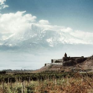 Un monastero in pietra si erge su una collina che domina un vasto vigneto, con una grande montagna innevata sullo sfondo sotto un cielo blu.