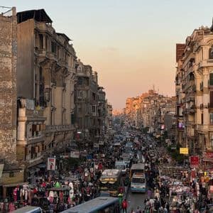 Una veduta dall'alto di una vivace strada cittadina affollata di persone, bancarelle, autobus e automobili tra alti edifici al tramonto.