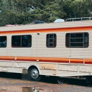 Una autocaravana grande, modelo antiguo, con rayas naranjas y rojas está estacionada en un terreno de grava junto a un bosque.