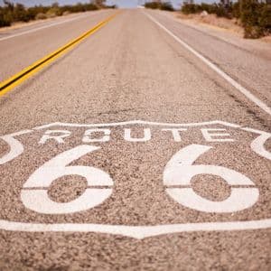 Un primer plano del cartel de la Ruta 66 pintado en una carretera de asfalto que se extiende a través de un paisaje seco y arbustivo.
