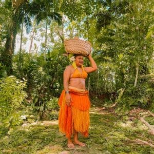 Una donna in abiti tradizionali arancioni trasporta un cesto intrecciato sulla testa mentre cammina su un sentiero in una giungla verde e lussureggiante.