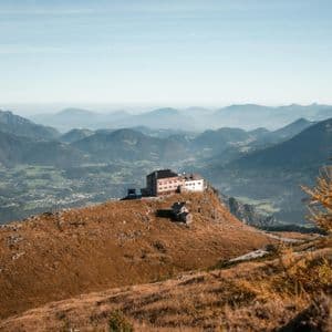 Ein großes Gebäude thront auf einem grasbewachsenen Berggipfel und überblickt ein weites Tal sowie geschichtete Bergketten unter einem klaren Himmel.