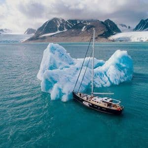 Una barca a vela naviga accanto a un grande iceberg su acqua turchese, con montagne innevate e un ghiacciaio sullo sfondo.