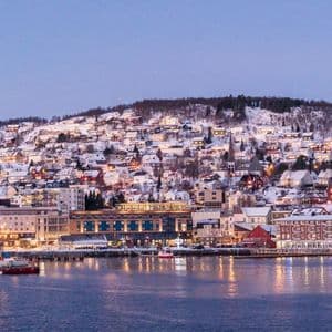 A snow-covered town built on a hillside along the water, with its buildings illuminated at dusk.