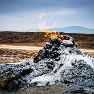 Un piccolo vulcano di fango erutta fiamme arancioni brillanti dal suo cratere in un paesaggio brullo sotto un cielo coperto.