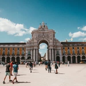 Folle di persone camminano attraverso una grande piazza assolata di fronte a un arco trionfale ornato sotto un cielo blu.