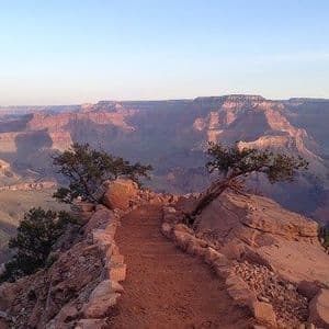 Un sentiero stretto e roccioso si snoda lungo il bordo di un vasto canyon con formazioni stratificate all'alba.