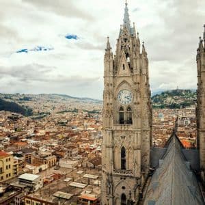 A tall, Gothic cathedral clock tower overlooking a cityscape that extends to distant hills under a cloudy sky.