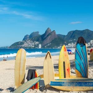Plusieurs planches de surf colorées se dressent sur le sable d'une plage animée, avec l'océan et les montagnes en arrière-plan.