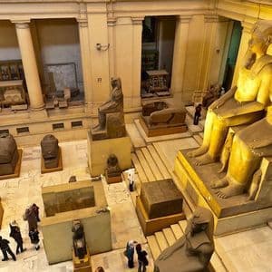 Una veduta dall'alto di una sala museale ricolma di colossali statue egizie antiche, sarcofagi e manufatti, con visitatori in basso.