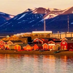 Case colorate di un villaggio di mare si riflettono nell'acqua calma, con grandi montagne innevate che si ergono sullo sfondo al tramonto.