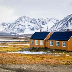 Due case di legno gialle con tetti scuri sorgono su un campo erboso e chiazzato di neve, di fronte a una vasta catena montuosa innevata.