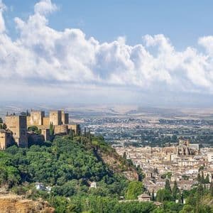 Una fortaleza histórica en una exuberante colina verde domina una ciudad en expansión y llanuras distantes bajo un cielo nublado.