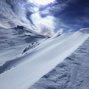 Un paisaje montañoso cubierto de nieve con una cresta barrida por el viento bajo un cielo azul profundo con nubes arremolinadas.