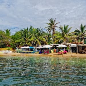 Spiaggia di sabbia con ristoranti colorati, palme e ombrelloni, vista dalla superficie dell'acqua.