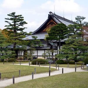 Un tradizionale edificio giapponese con tetto in tegole grigie si staglia dietro un giardino curato, con un sentiero di ghiaia serpeggiante e vari alberi.