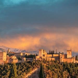 Una fortaleza de piedra histórica en una colina boscosa es iluminada por un atardecer dorado, con montañas visibles al fondo.
