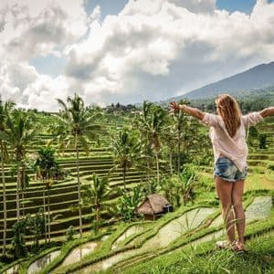 Eine Frau steht mit dem Rücken zur Kamera auf einem grasbewachsenen Hügel, die Arme ausgestreckt, mit Blick auf weite Reisterrassen und Palmen.