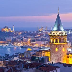 Una torre di pietra illuminata con tetto conico domina un vasto skyline cittadino con moschee sull'acqua al tramonto.