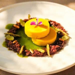 Un piatto gourmet su un piatto bianco con due budini gialli sovrapposti su grani rossi e una salsa verde, guarnito con un fiore.