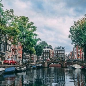 Une vue sur un canal urbain bordé de bâtiments traditionnels, d'arbres verts et de bateaux amarrés, avec un pont de pierre au loin.
