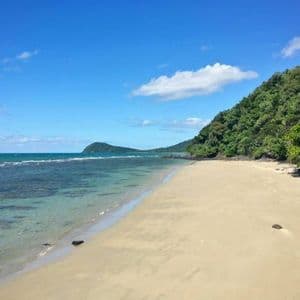 Una playa de arena se extiende junto a aguas cristalinas de color turquesa y una exuberante colina verde bajo un cielo azul con nubes dispersas.