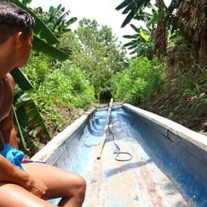 Une personne est assise dans un long bateau bleu, naviguant sur un canal étroit entouré d'une végétation luxuriante de jungle et de bananiers.