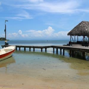 Un bateau rouge et blanc est amarré dans des eaux claires et peu profondes, à côté d'une jetée en bois avec une cabane au toit de chaume sous un ciel partiellement nuageux.