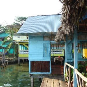 Des maisons colorées en bois sur pilotis se dressent sur l'eau dans un village tropical avec des passerelles en bois.